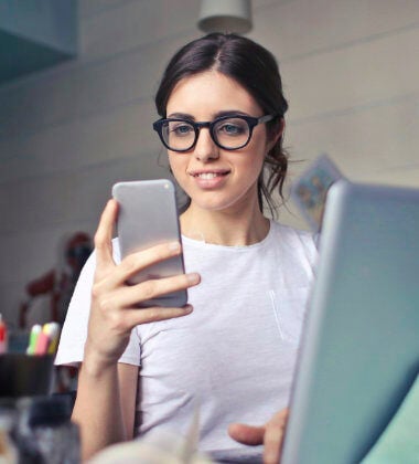 A young woman using a smartphone whilst sitting in front of a laptop